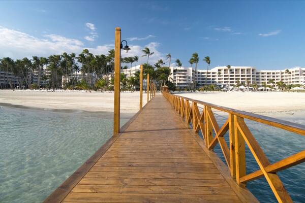 Un muelle de madera que lleva a una playa con agua turquesa cristalina, palmeras y edificios de un resort al fondo.