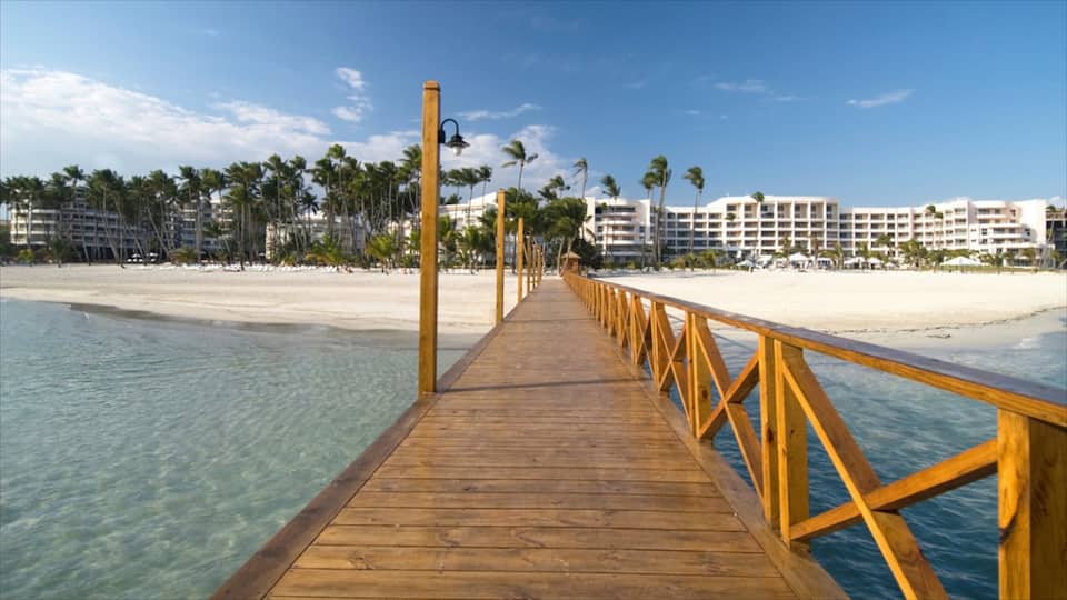A wooden pier leading to a beach with clear turquoise water, palm trees, and resort buildings in the background.