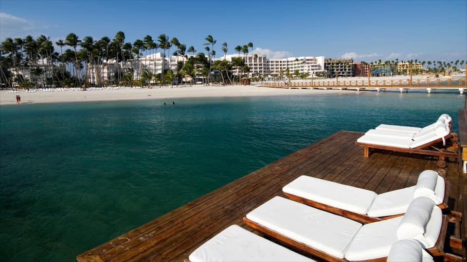 A wooden deck with white lounge chairs overlooking turquoise waters, palm trees, and a beach with hotel buildings in the background.