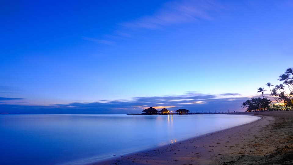 A beach at dusk with calm waters, overwater bungalows, and palm trees.