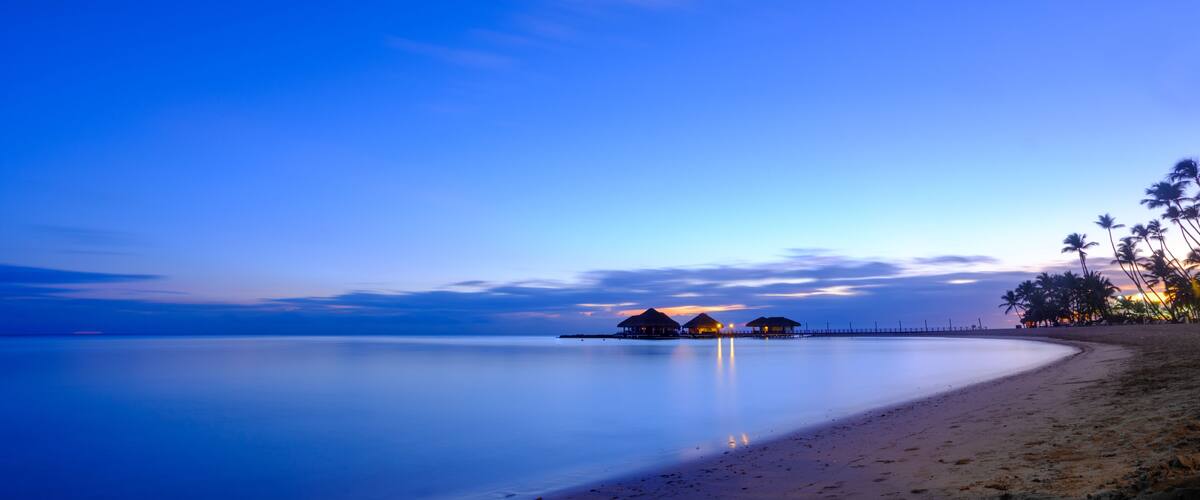 A beach at dusk with calm waters, overwater bungalows, and palm trees.