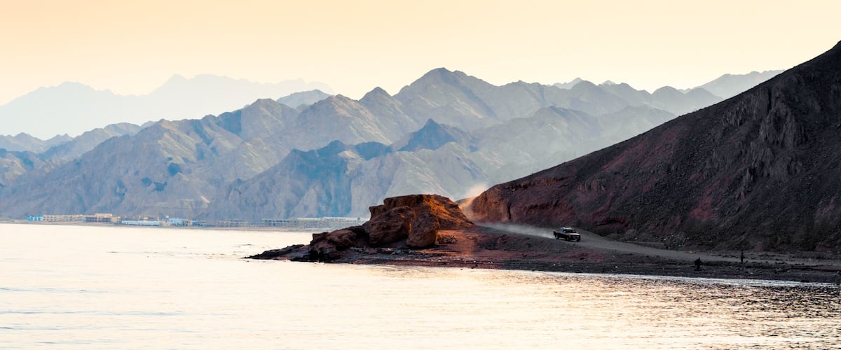 A rugged coastline with a rocky outcrop, a vehicle driving along a dirt path, and a mountain range in the background.