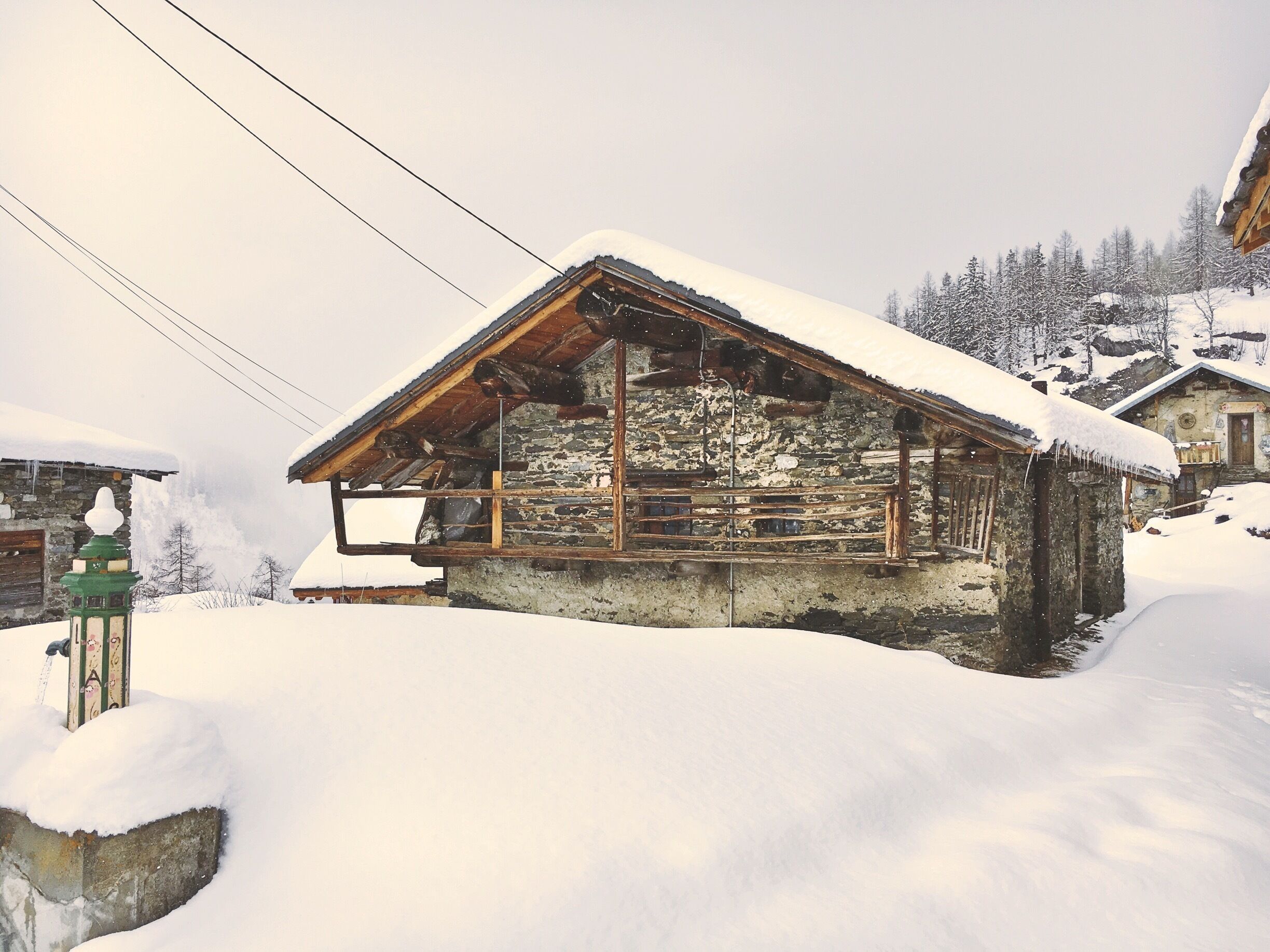 A snow-covered wooden house with a stone foundation, surrounded by snow-covered trees and a mountainous backdrop.