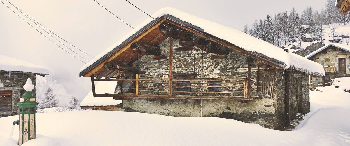 A snow-covered wooden house with a stone foundation, surrounded by snow-covered trees and a mountainous backdrop.
