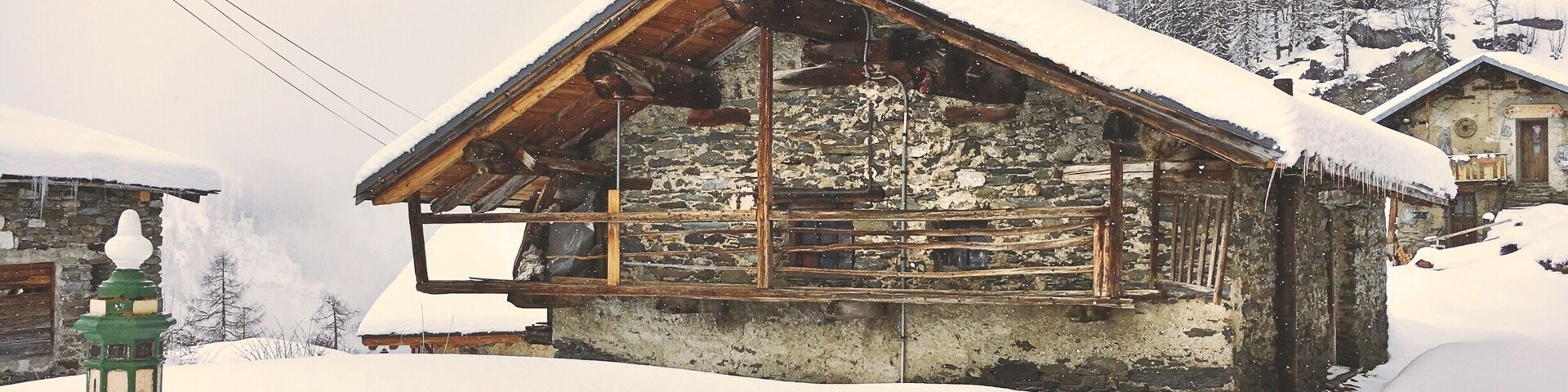 A snow-covered wooden house with a stone foundation, surrounded by snow-covered trees and a mountainous backdrop.