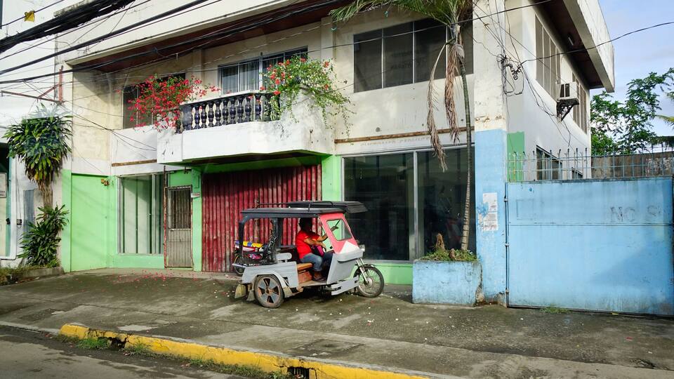 A tuk-tuk parked in front of a building with a red gate and a balcony with red flowers.