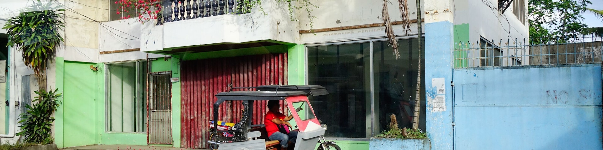 Un tuk-tuk estacionado frente a un edificio con una puerta roja y un balcón con flores rojas.