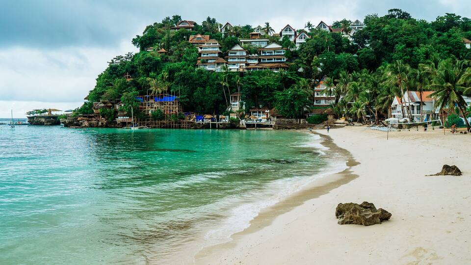 A beach with clear turquoise water, a sandy shore, and a hillside with buildings and palm trees.