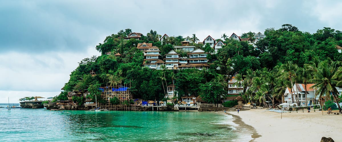 A beach with clear turquoise water, a sandy shore, and a hillside with buildings and palm trees.