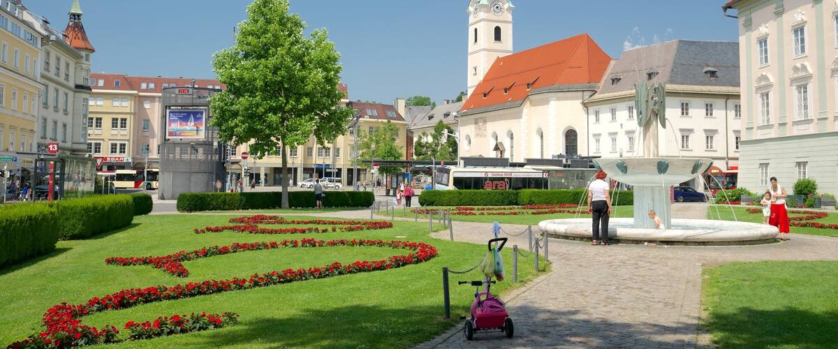A public square with a fountain, red flower beds, and historic buildings.