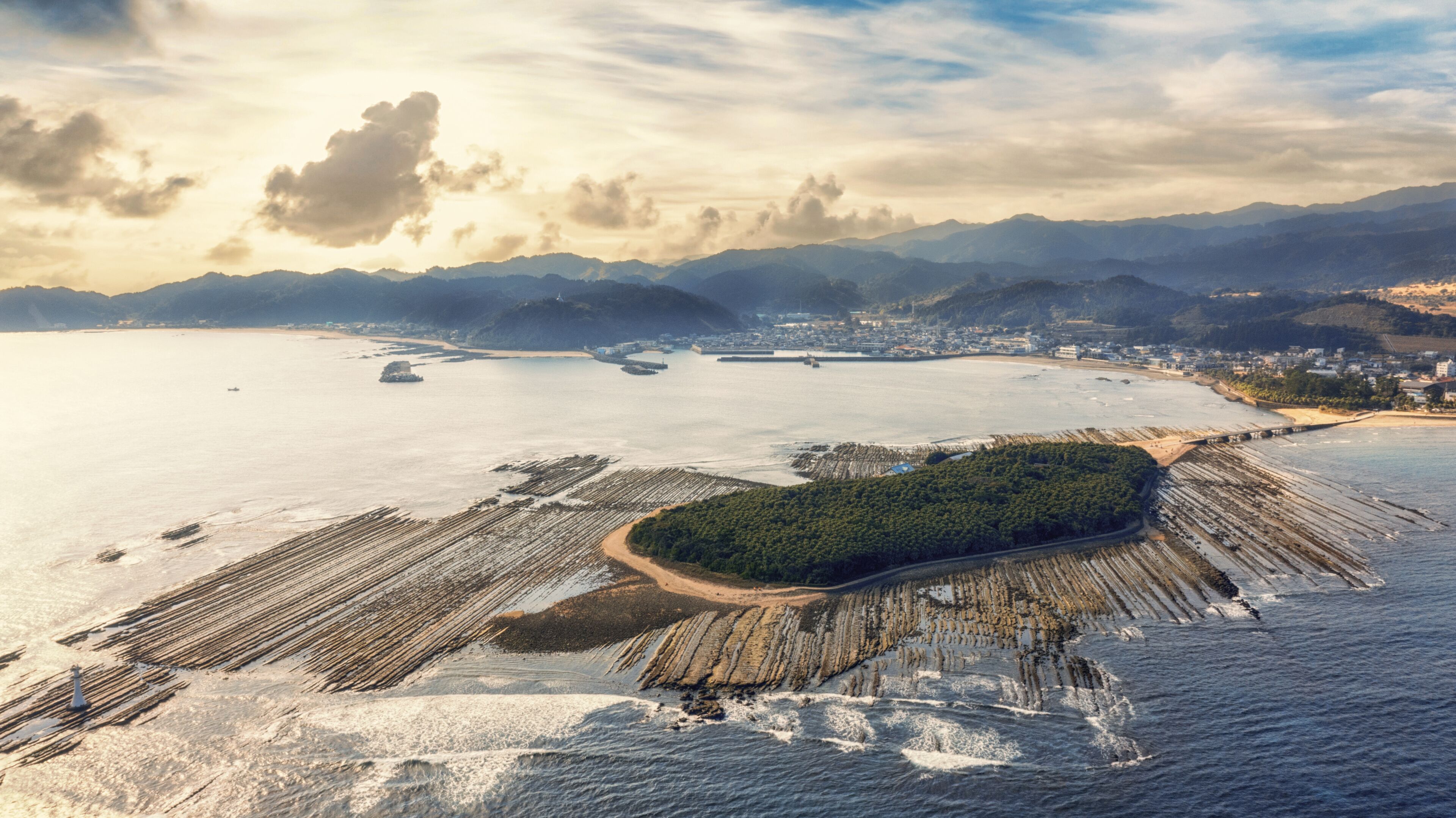 An aerial view of a coastal area with a forested island, a sandy beach, and a marina with boats.