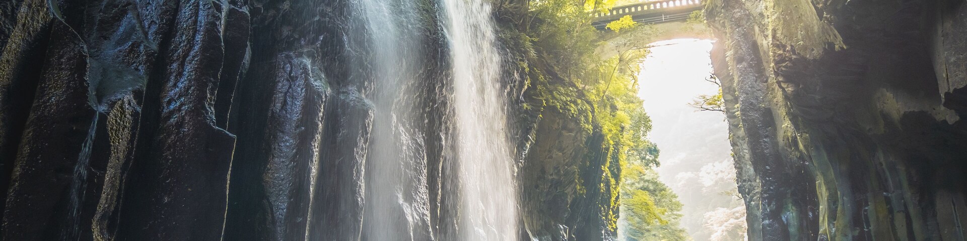 Una cascada se precipita en una piscina turquesa, rodeada de acantilados rocosos y un puente al fondo.