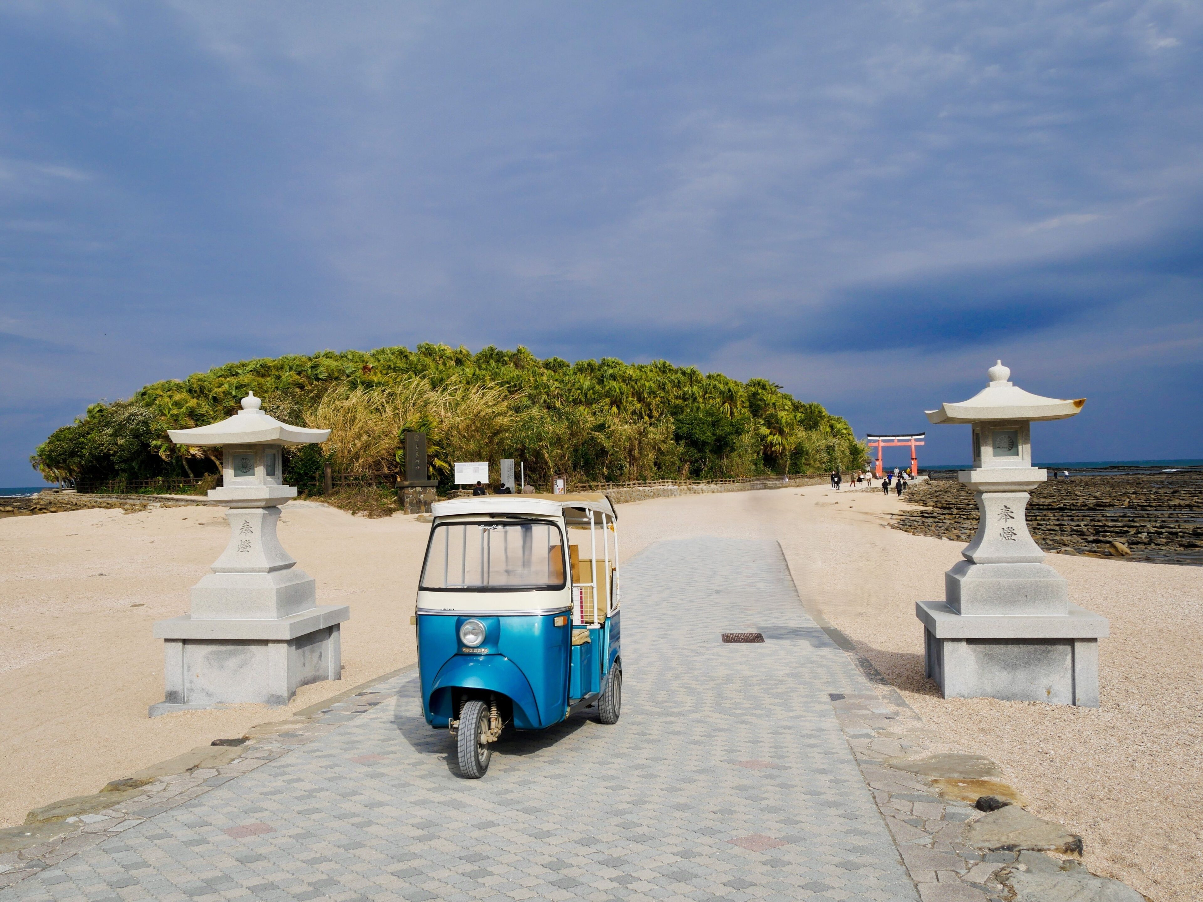 A blue tuk-tuk on a paved path leading to a beach with palm trees and a red torii gate in the background.