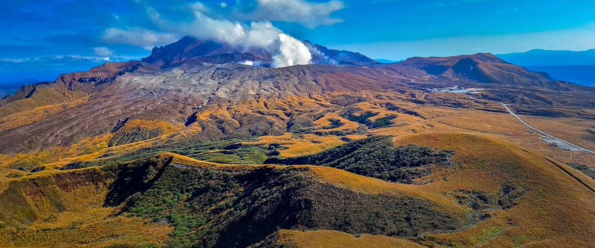 A volcanic mountain with smoke, a winding road, and rolling hills.