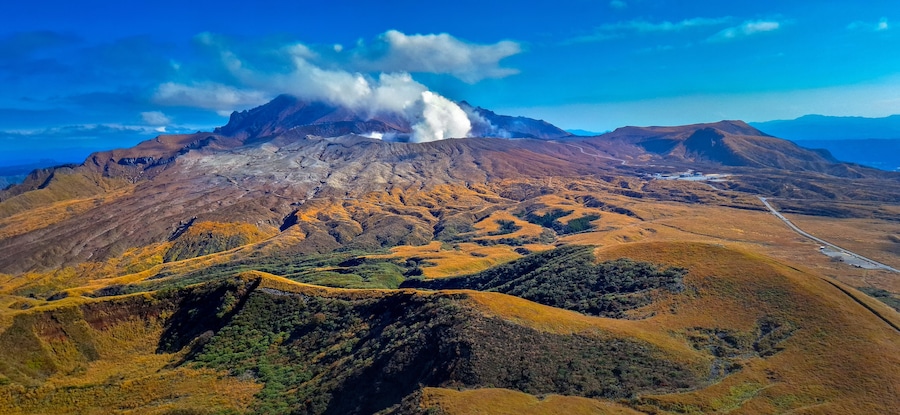 A volcanic mountain with smoke, a winding road, and rolling hills.