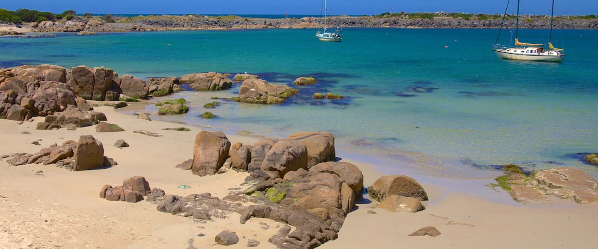 A rocky beach with clear turquoise water and sailboats.