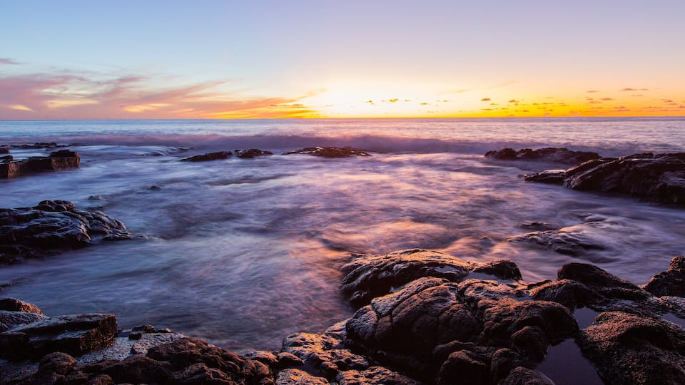 A sunset over a rocky coastline with waves crashing against the rocks.