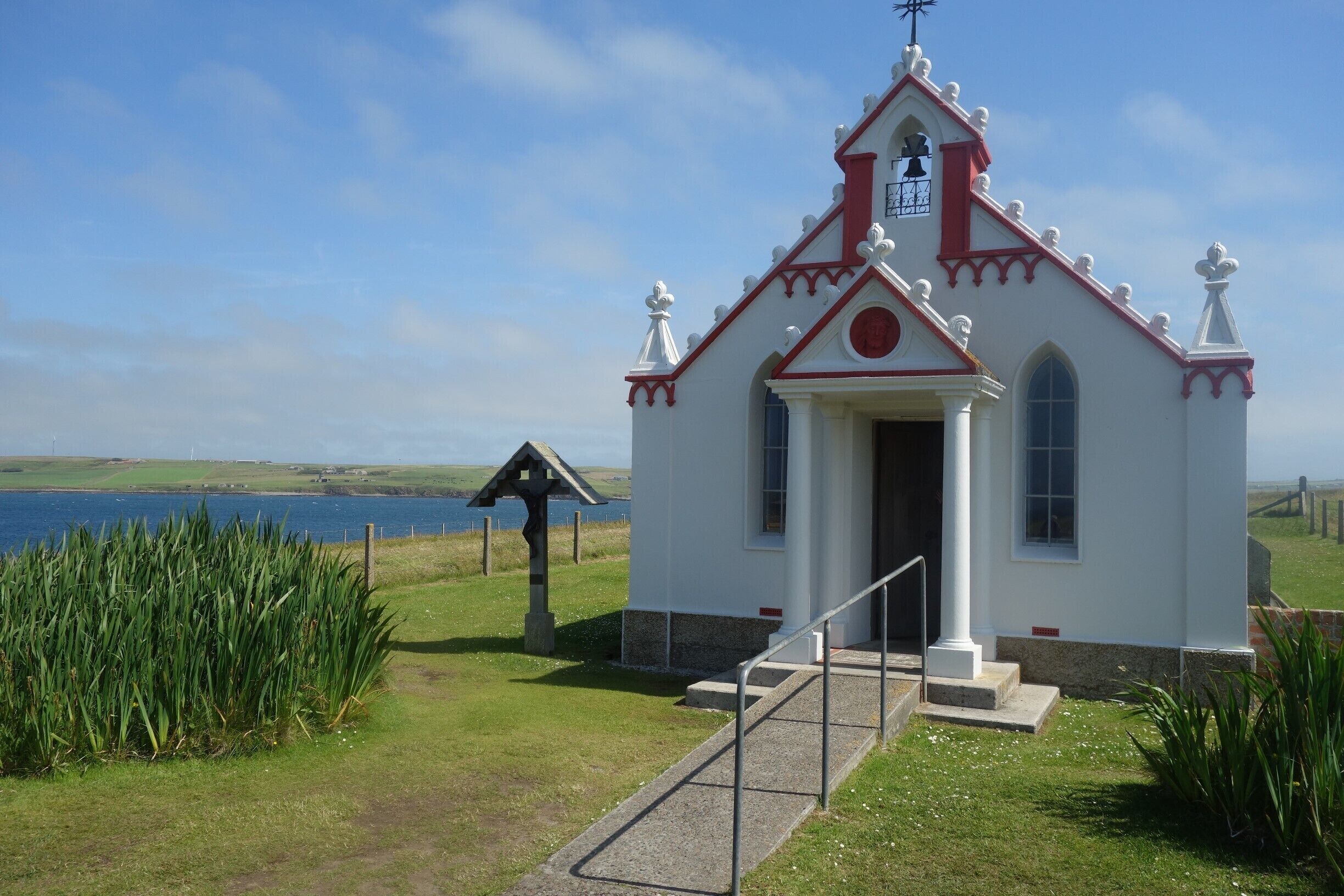 A small white church with a red roof and a bell tower, located near a body of water.