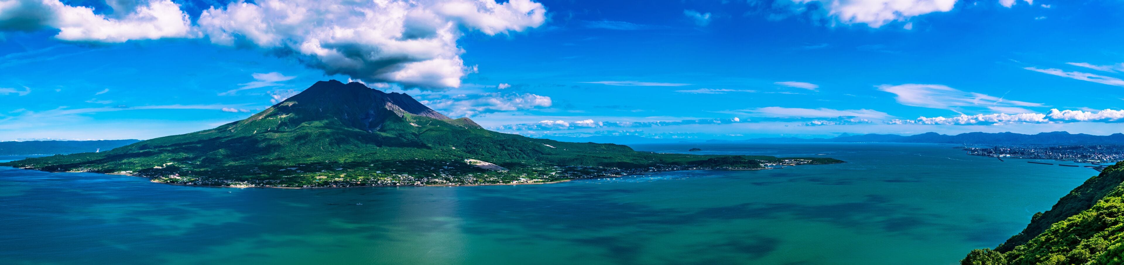 Una isla volcánica con un pico destacado, rodeada de aguas azul turquesa y una ciudad a lo lejos.