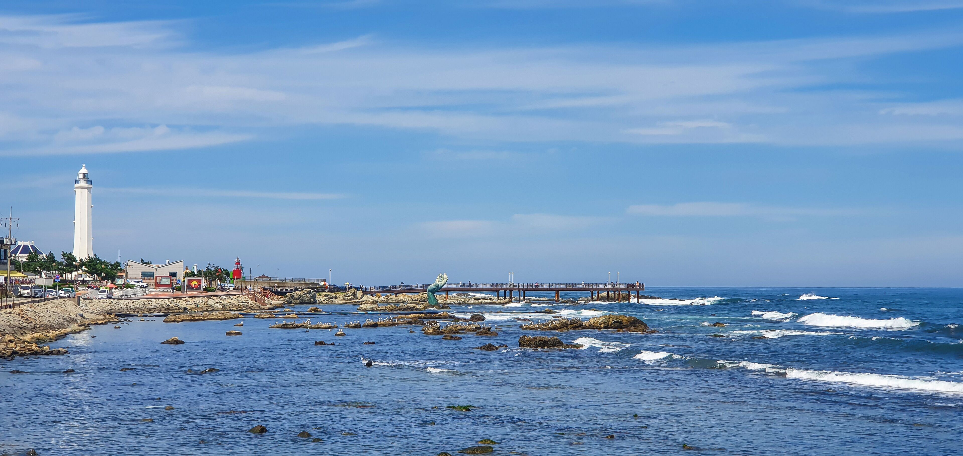 A coastal scene with a lighthouse, rocky shoreline, waves, and a pier.