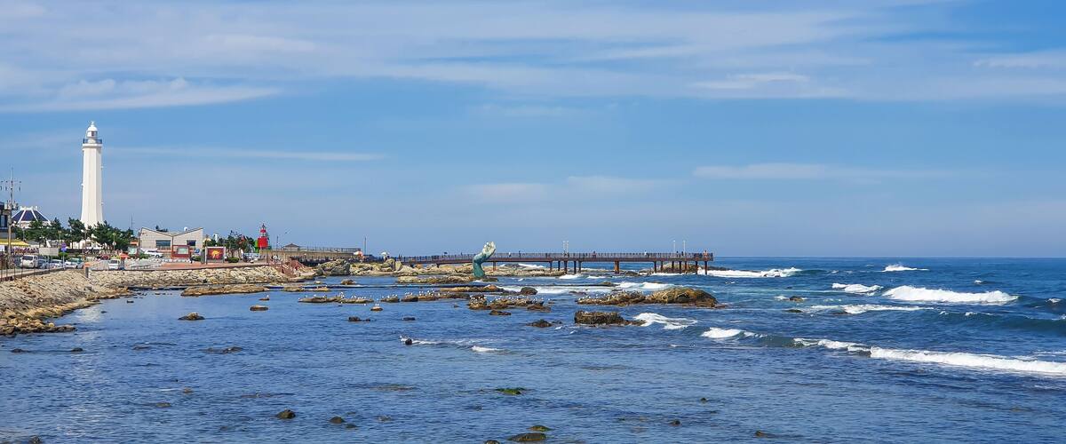 A coastal scene with a lighthouse, rocky shoreline, waves, and a pier.