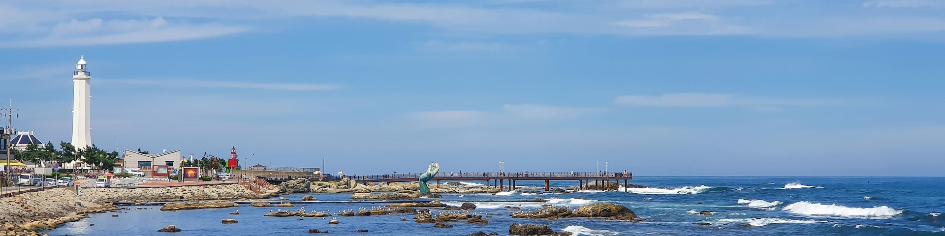 A coastal scene with a lighthouse, rocky shoreline, waves, and a pier.