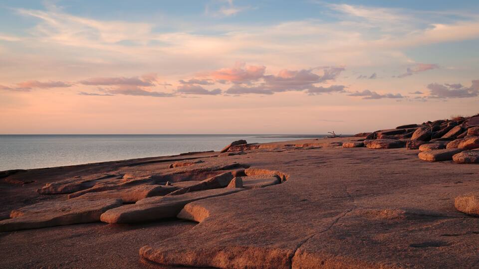 A rocky shoreline with large boulders and a calm sea under a sunset sky.