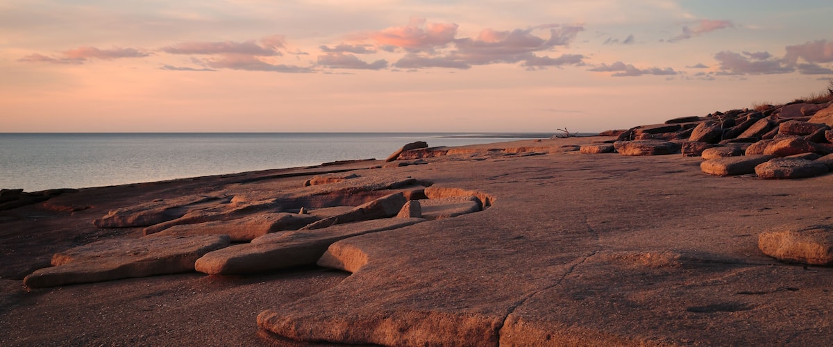 A rocky shoreline with large boulders and a calm sea under a sunset sky.