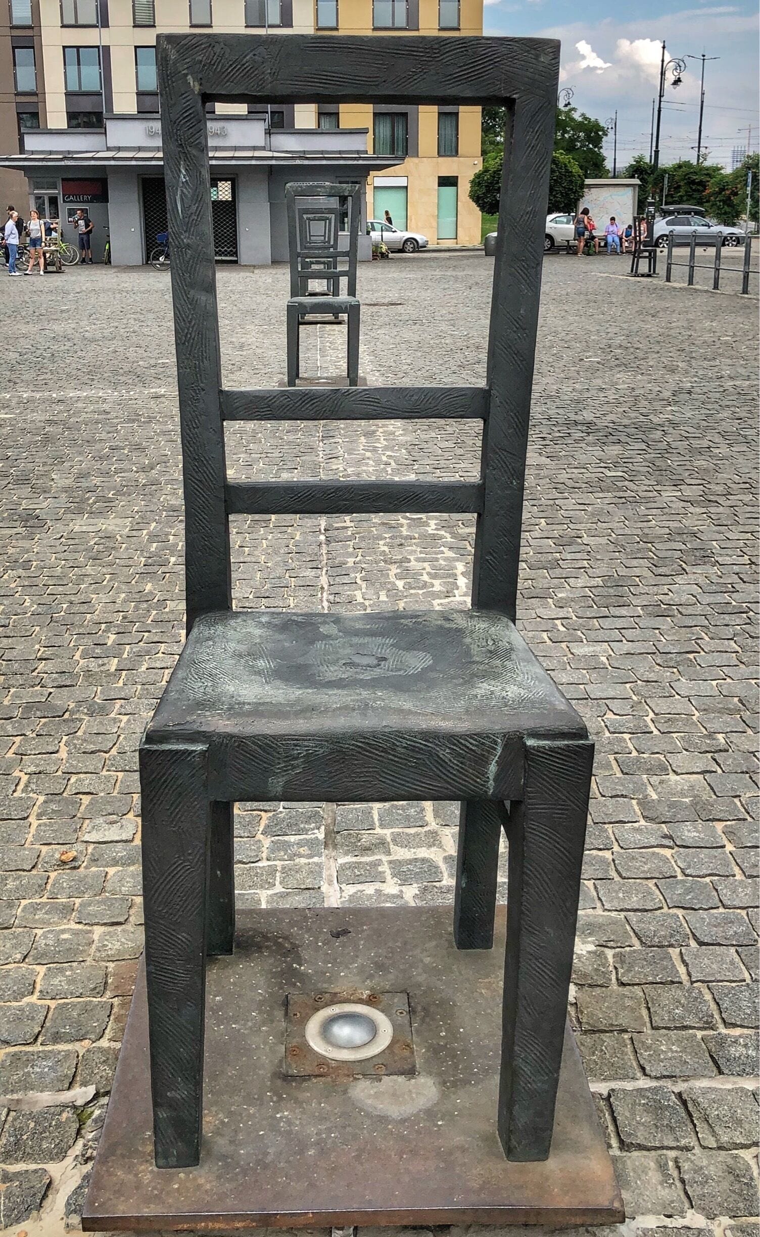 A metal chair with a square base and a backrest, placed on a cobblestone street.