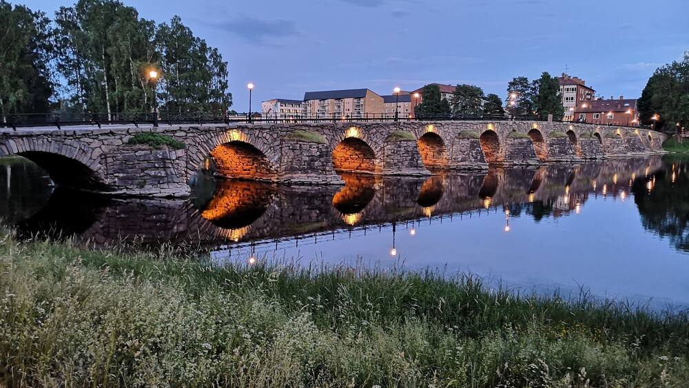 Un pont de pierre enjambant une rivière paisible au crépuscule, les lumières se reflétant sur l’eau.