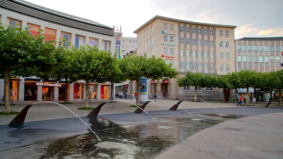A public square with a water feature, trees, and buildings with commercial establishments.