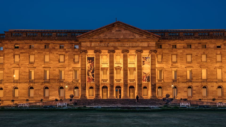 A large, illuminated building with classical architecture, including columns and a central pediment, at dusk.