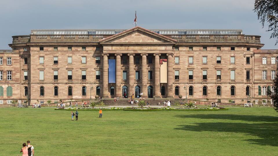 A large, historic building with a central entrance and columns, surrounded by a well-maintained lawn and trees.