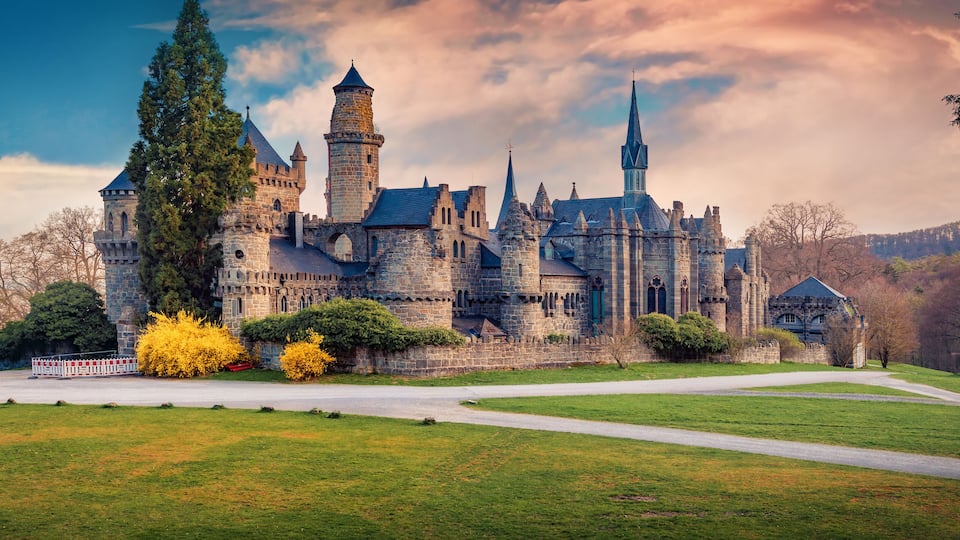A large castle with multiple towers and turrets, surrounded by a well-maintained lawn and trees.