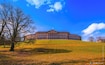 A large, historic building with a central dome and multiple wings, surrounded by a grassy hill and leafless trees.