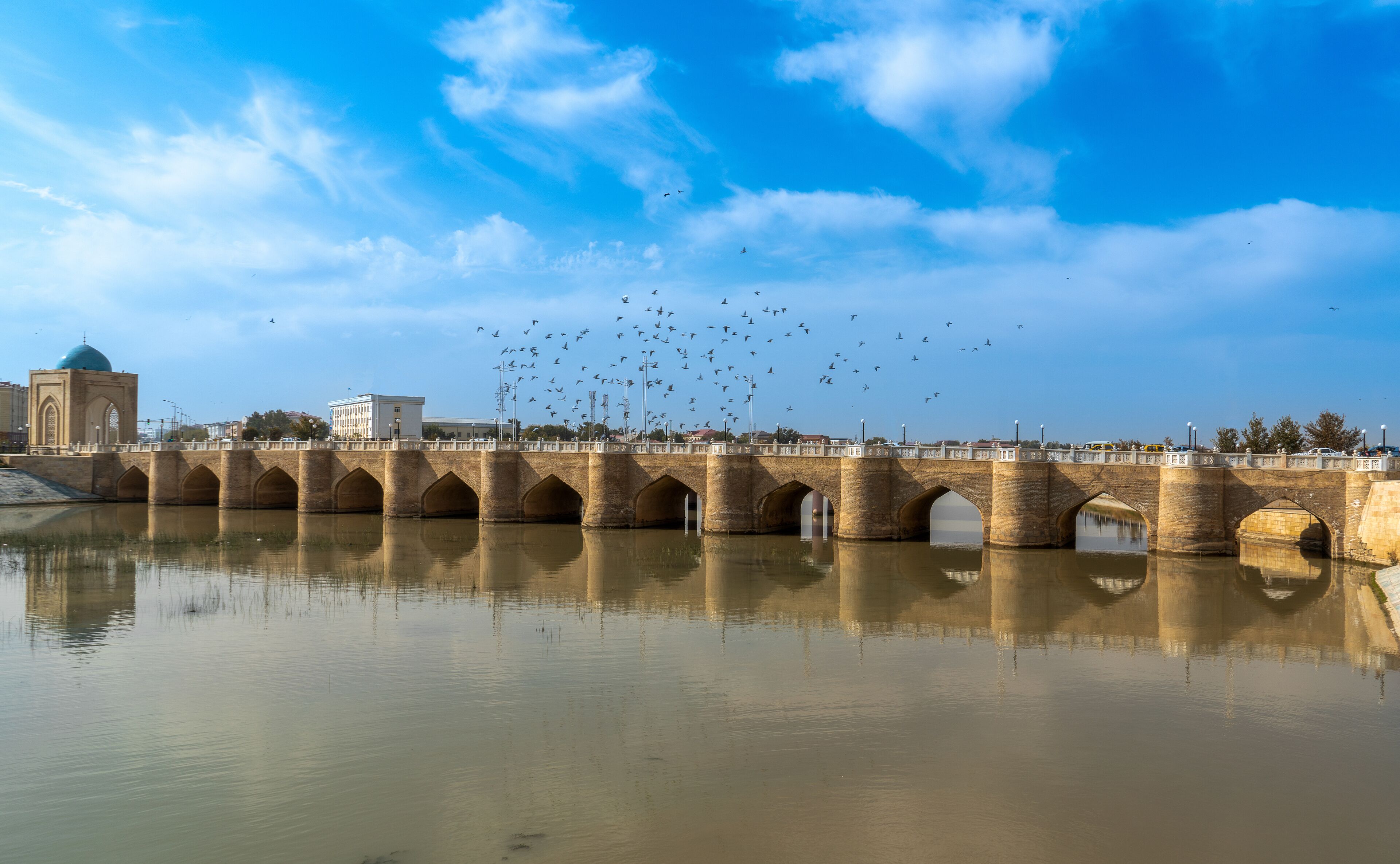 A bridge with multiple arches over a calm river, a mosque with a blue dome, and a flock of birds flying in the sky.
