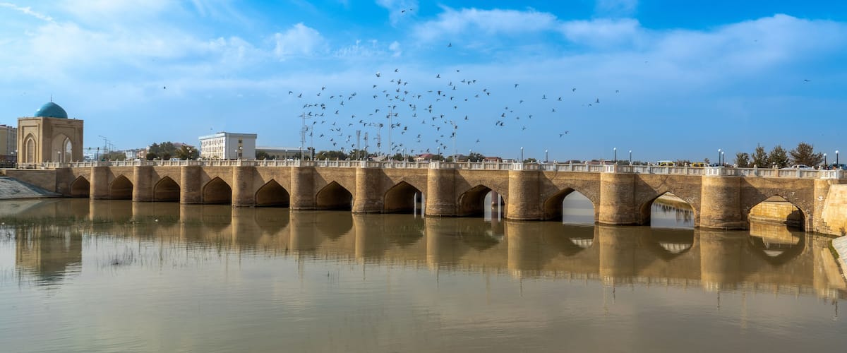A bridge with multiple arches over a calm river, a mosque with a blue dome, and a flock of birds flying in the sky.