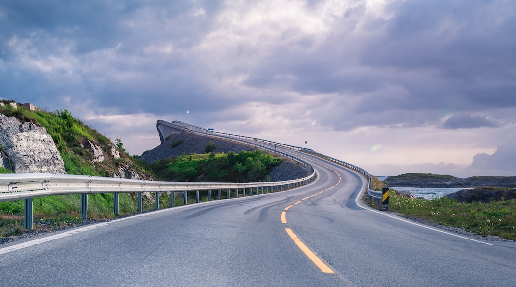 A curving road with guardrails, a mountainous landscape, and a body of water in the distance.