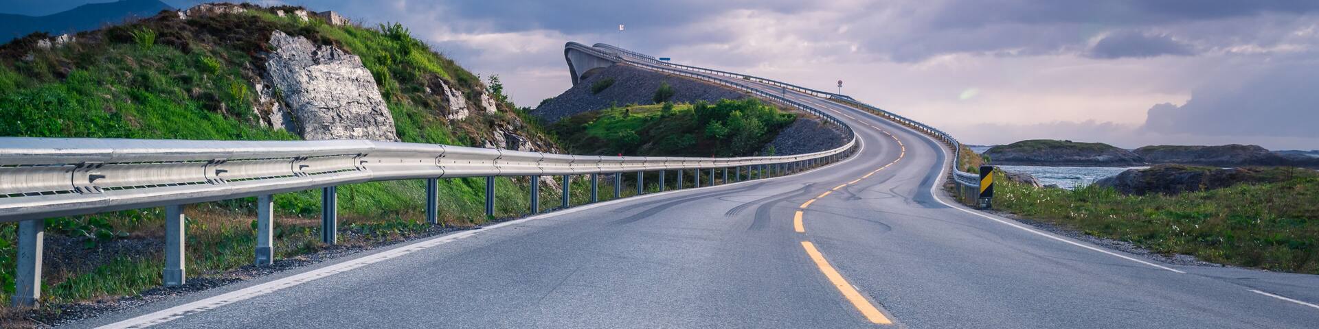 A curving road with guardrails, a mountainous landscape, and a body of water in the distance.
