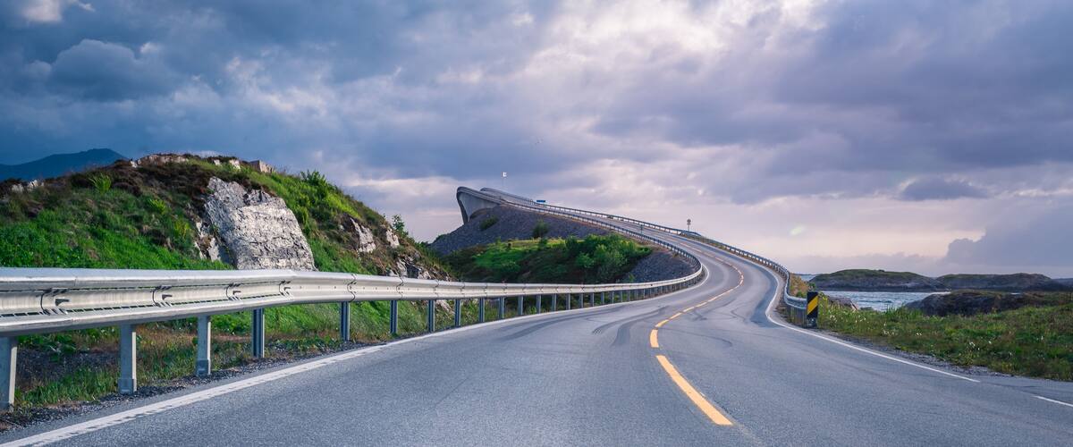 A curving road with guardrails, a mountainous landscape, and a body of water in the distance.