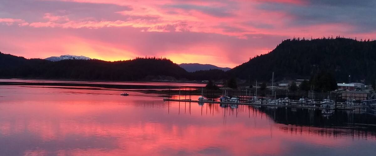 A calm lake at sunset with a reflection of the sky, surrounded by hills and a marina with boats.