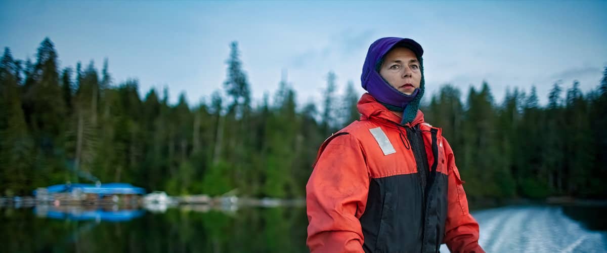 A person in a red and black jacket standing on a boat with a forest and water in the background.