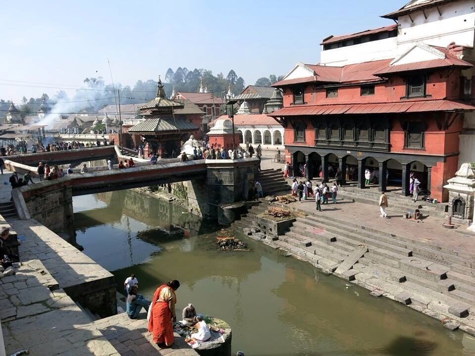 A riverside temple complex with a bridge, steps leading into the water, and people around.