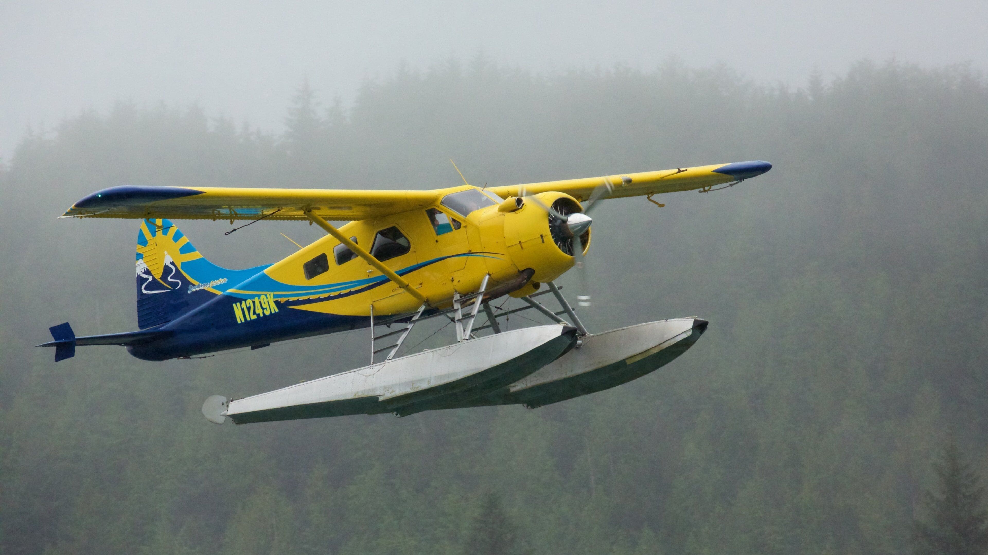 A yellow and blue seaplane with the registration N1249K flying over a foggy forested area.