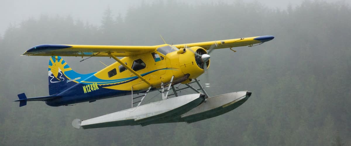 A yellow and blue seaplane with the registration N1249K flying over a foggy forested area.