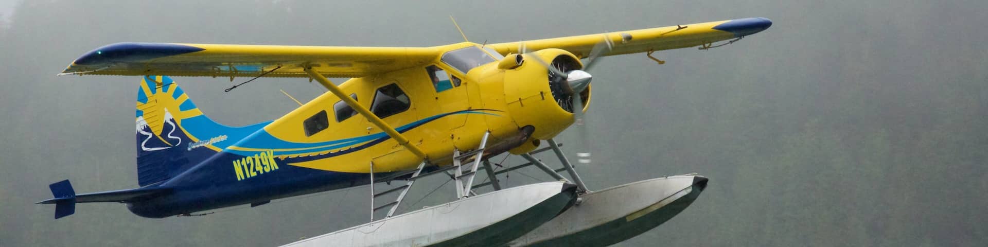 A yellow and blue seaplane with the registration N1249K flying over a foggy forested area.
