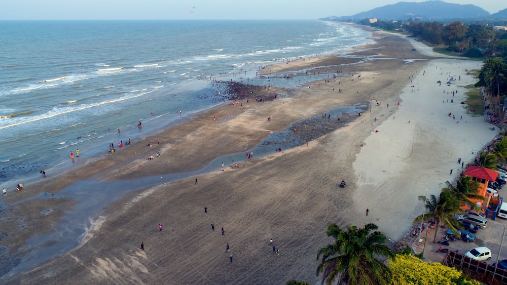 Une vaste plage où l’on voit des gens se promener et nager, des palmiers et, au loin, une chaîne de montagnes.
