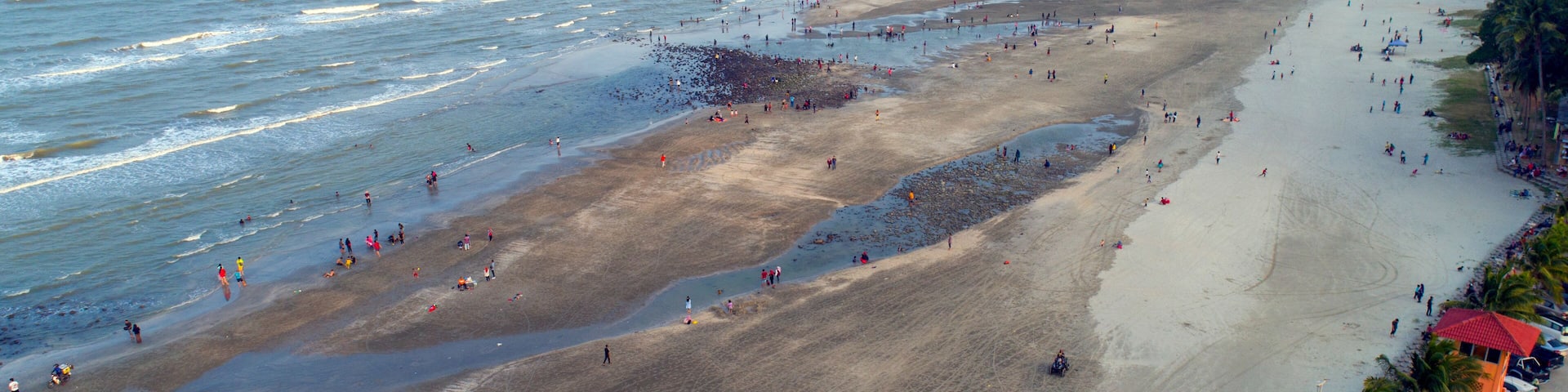 A wide beach with people walking and swimming, palm trees, and a distant mountain range.