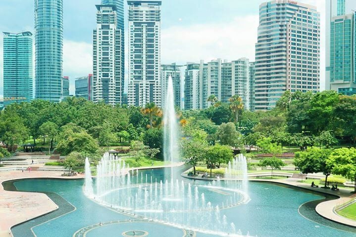 A city park with a central fountain, surrounded by trees and modern high-rise buildings.