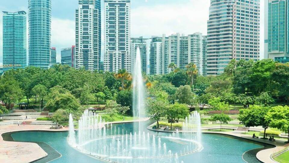 A city park with a central fountain, surrounded by trees and modern high-rise buildings.